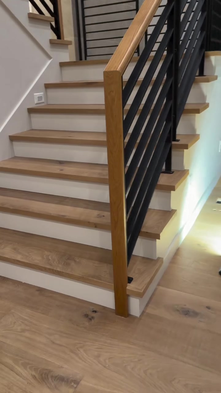 A white oak staircase with black metal railing — treads built from the floor material, flowing into the wide-plank floor below